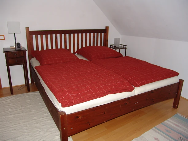 Ferienwohnung Am historischen Rathaus - Schlafzimmer Schlafzimmer mit einem Holzbett, roten Bettdecken, Nachttisch und heller, schräger Decke.