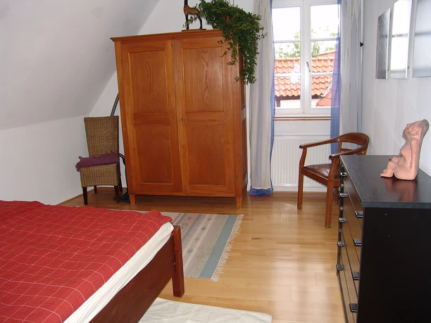 Ferienwohung Am Historischen Rathaus - Schlafzimmer Ein helles Schlafzimmer mit Holzboden, Bett, Holzschrank, Sessel und Fenstern mit Blick aufs Dach.