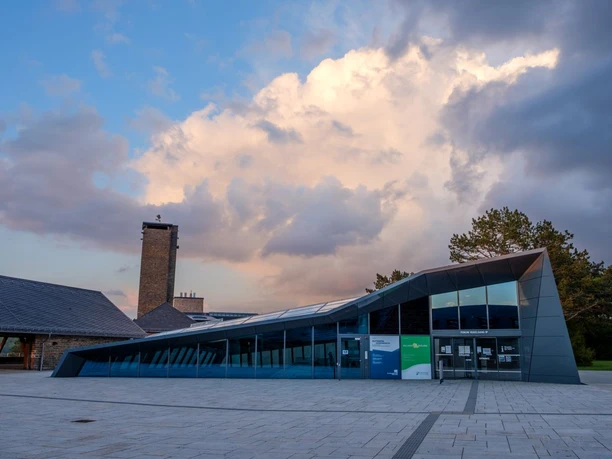 Adlerhof Vogelsang IP Modernes, abflachendes Gebäude mit Glasfront auf einem gepflasterten Platz bei Dämmerung.