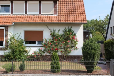 FeWo Pohlmann Ferienwohnung in einem Haus mit roten Dachziegeln, umgeben von gepflegtem Garten und Rosenstrauch.