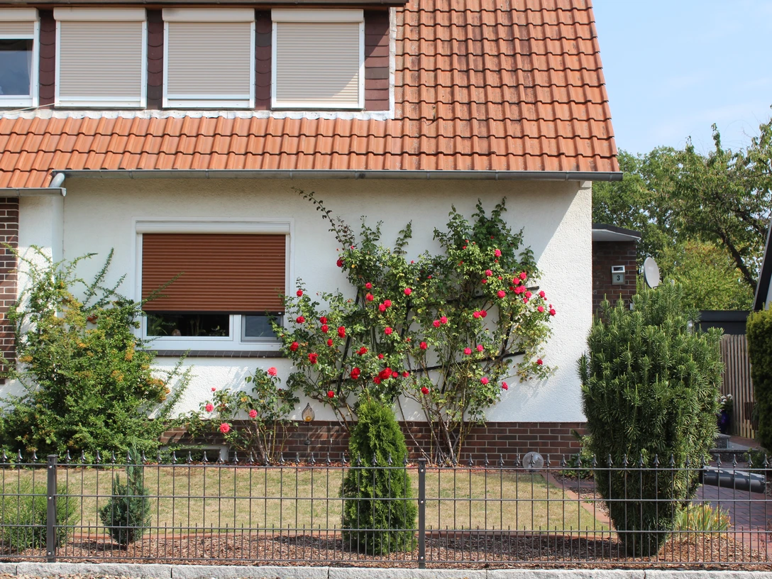 FeWo Pohlmann Ferienwohnung in einem Haus mit roten Dachziegeln, umgeben von gepflegtem Garten und Rosenstrauch.