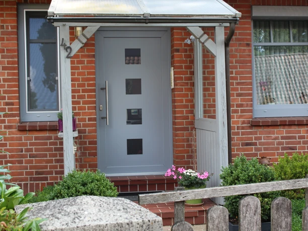 Entrance to a vacation apartment with glazed canopy, modern gray door, floral decor and brick construction.