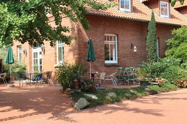 Gebouw van rode baksteen met terras, tuinmeubilair en groene planten onder blauwe parasols.