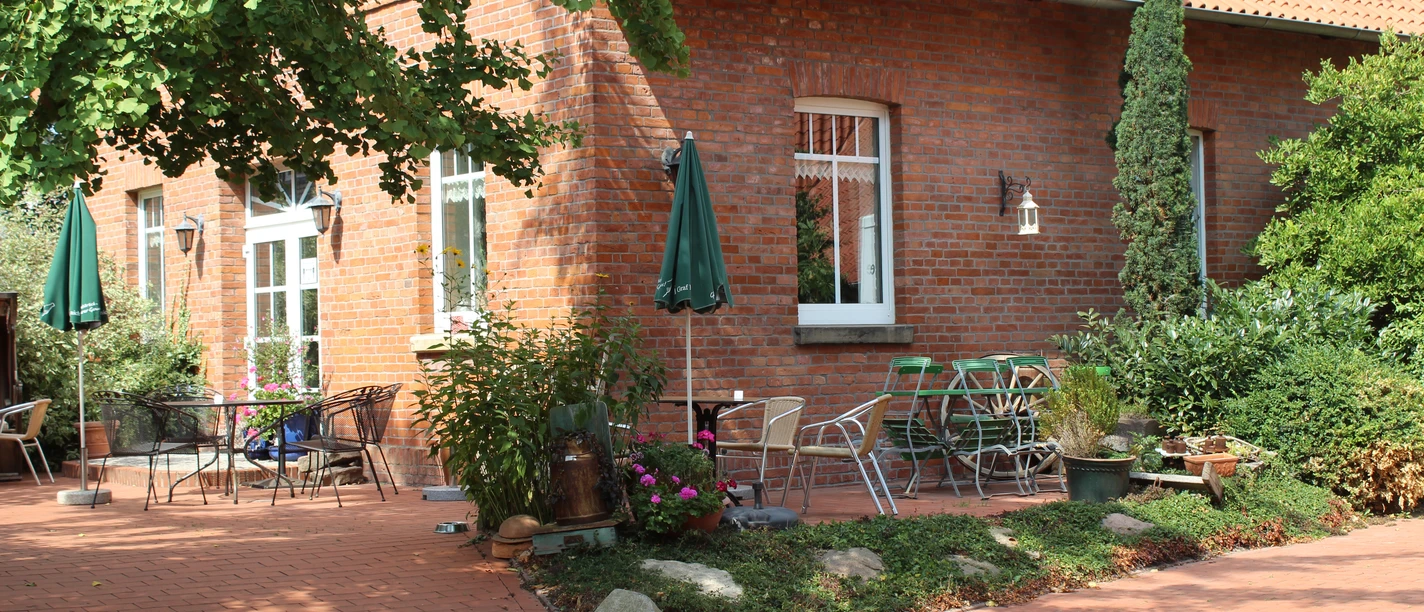 Gebouw van rode baksteen met terras, tuinmeubilair en groene planten onder blauwe parasols.
