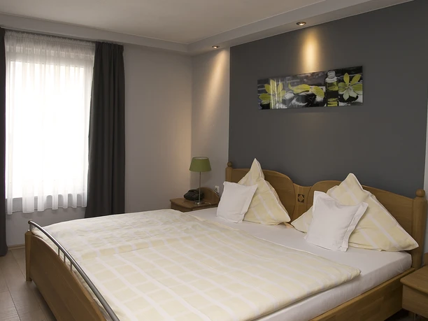 Doppelzimmer Doppelzimmer mit Holzbetten, zwei Kissen und Lampe, vor einer grauen Wand mit Blumenbild.