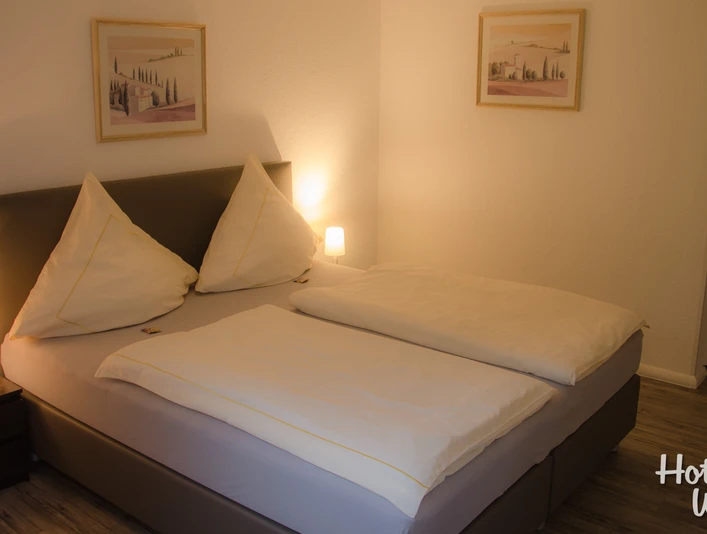 Doppelzimmer Ein einladendes Doppelzimmer mit zwei Nachttischen, Gemälden an den Wänden und stehendem Holzfußboden.