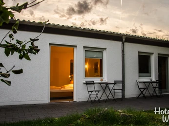 Terasse Ein Haus mit Flachdach, drei offenen Türen, Abendstimmung und bereitstehenden Terrassenmöbeln.