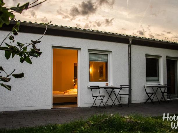 Terasse Ein Haus mit Flachdach, drei offenen Türen, Abendstimmung und bereitstehenden Terrassenmöbeln.