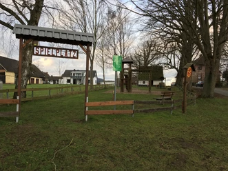 Kinderspielplatz Biesterfeld mit Holztor, Klettergerüst und Bänken unter Bäumen an einer ruhigen Straße.