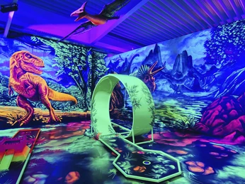 Blacklight_District2_foto_ccbysa_foto©adventure-district.jpeg Das Bild zeigt einen Dinosaurier-Themen-Minigolfplatz im Neonlicht, umgeben von Wandgemälden prähistorischer Tiere und Pflanzen.