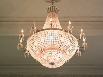 spiegelsaal-kronleuchter.jpg Ein prächtiger Kronleuchter aus Kristallglas beleuchtet elegant einen kunstvoll verzierten Raum mit Stuckdecke.
