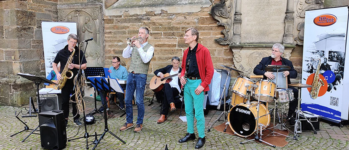 jazzgroup792x356.jpg Eine sechsköpfige Jazzband spielt im Freien vor einer historischen Steinwand auf einer gepflasterten Fläche.