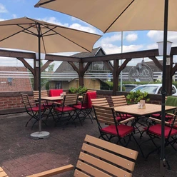 NieheimRestaurantDakos.jpg Sonnige Außenterrasse mit Holztischen und roten Stühlen, umgeben von Holzzaun und Sonnenschirmen.