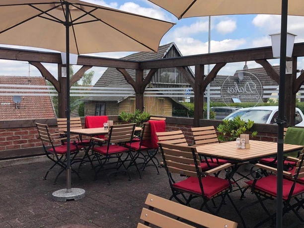 NieheimRestaurantDakos.jpg Sonnige Außenterrasse mit Holztischen und roten Stühlen, umgeben von Holzzaun und Sonnenschirmen.