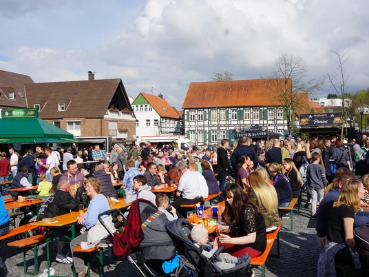 Viele Menschen sitzen und stehen an Bierbänken auf einem belebten Marktplatz unter freiem Himmel.