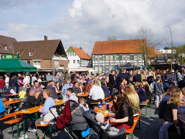 Viele Menschen sitzen und stehen an Bierbänken auf einem belebten Marktplatz unter freiem Himmel.