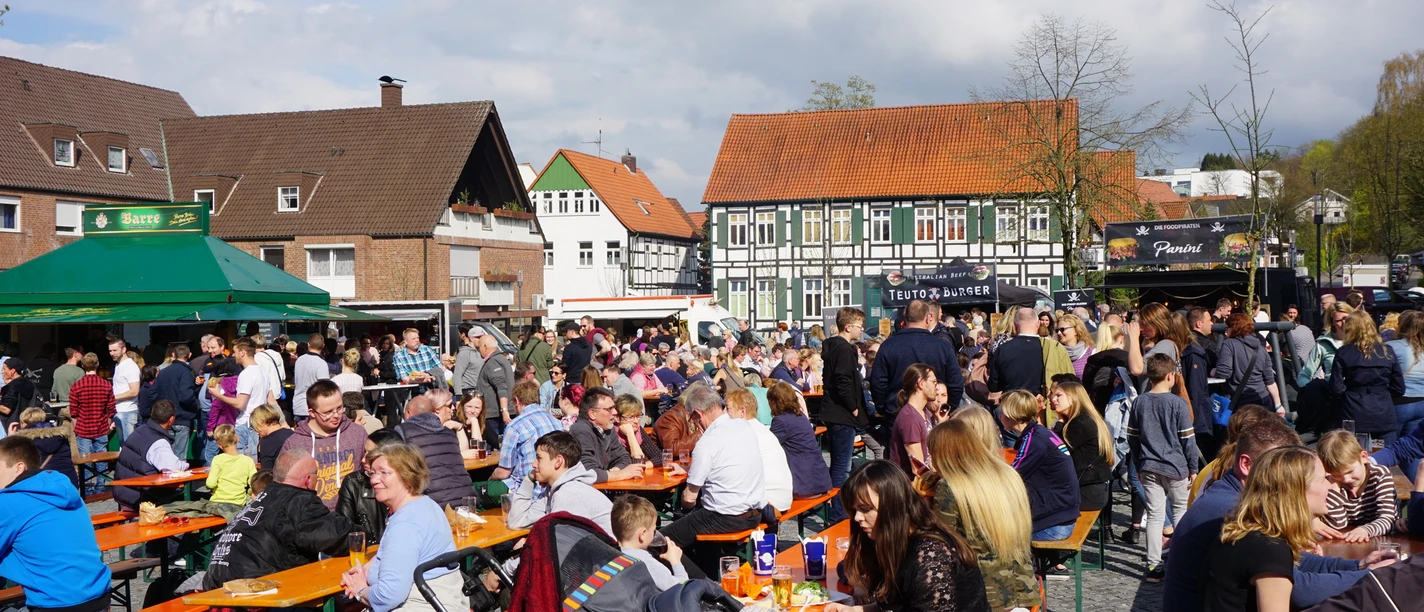 Viele Menschen sitzen und stehen an Bierbänken auf einem belebten Marktplatz unter freiem Himmel.