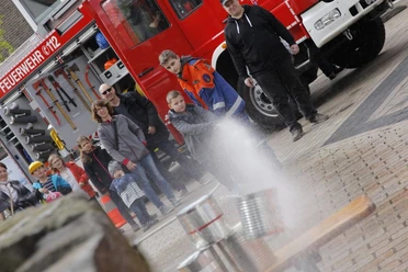 Kinder und Erwachsene bei einer Feuerwehrveranstaltung sehen einem jungen Teilnehmer zu, der mit einem Schlauch spritzt.