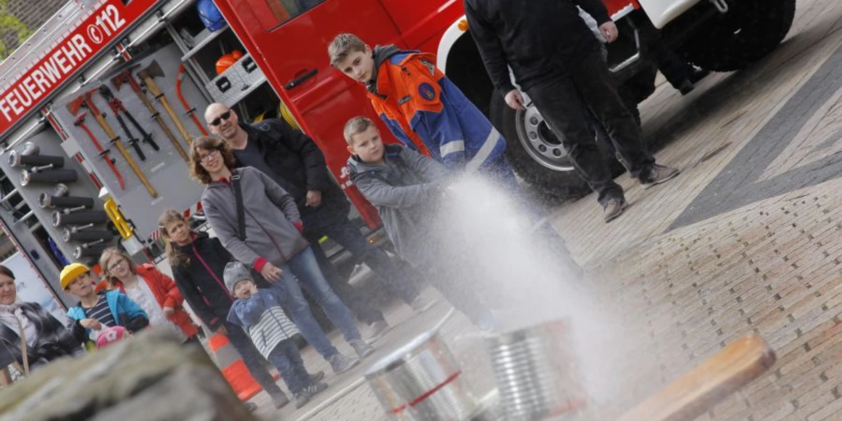 kinderfest2_jugendfeuerwehr.jpg Kinder und Erwachsene bei einer Feuerwehrveranstaltung sehen einem jungen Teilnehmer zu, der mit einem Schlauch spritzt.