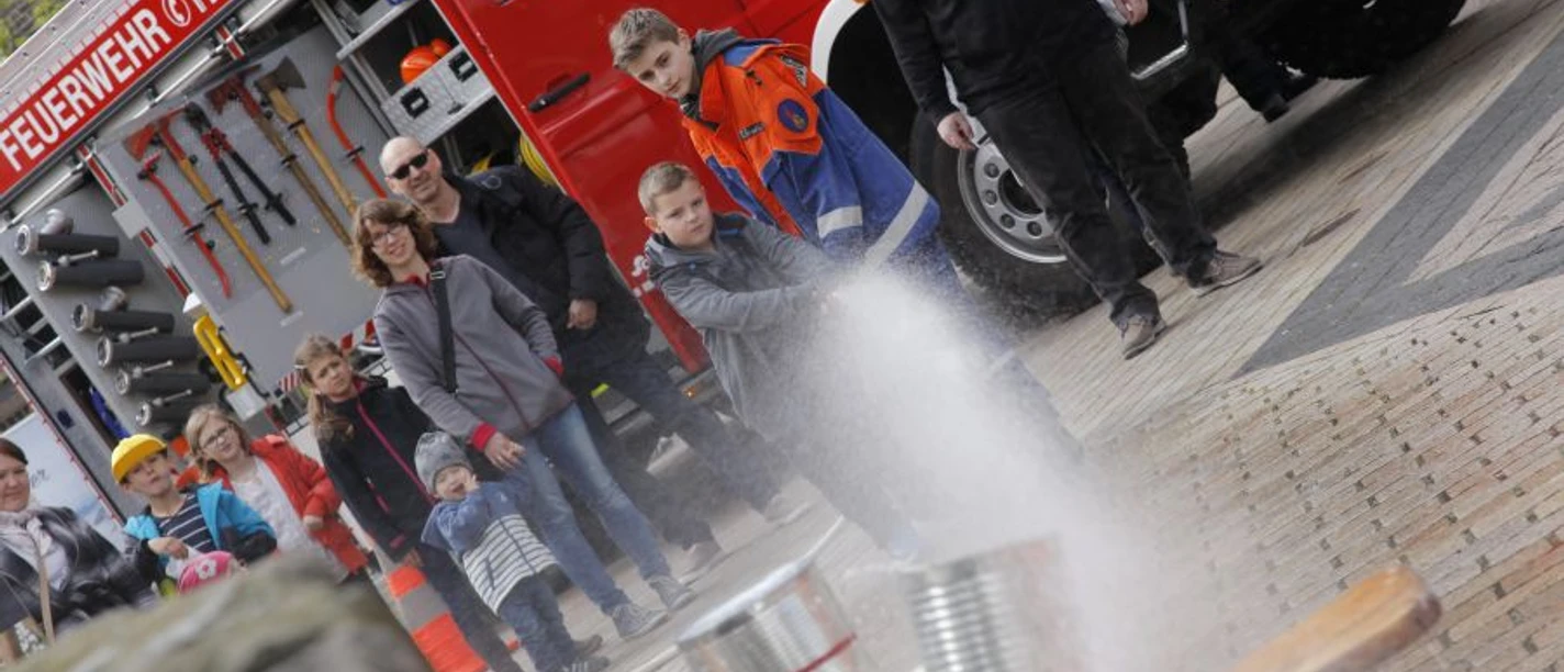kinderfest2_jugendfeuerwehr.jpg Kinder und Erwachsene bei einer Feuerwehrveranstaltung sehen einem jungen Teilnehmer zu, der mit einem Schlauch spritzt.