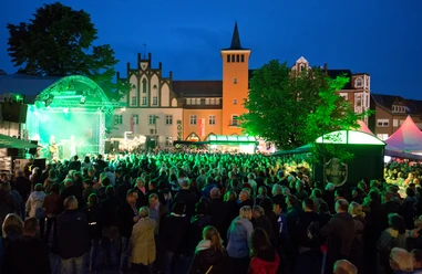Lübbecke_Bierbrunnenfest1_Oliver Krato.jpg Abendliches Festival mit beleuchteter Bühne und Menschenmenge auf einem Marktplatz in Lübbecke.