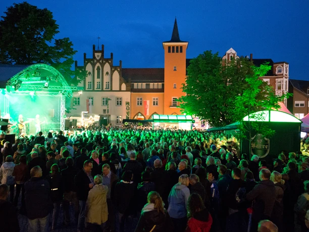 Lübbecke_Bierbrunnenfest1_Oliver Krato.jpg Abendliches Festival mit beleuchteter Bühne und Menschenmenge auf einem Marktplatz in Lübbecke.