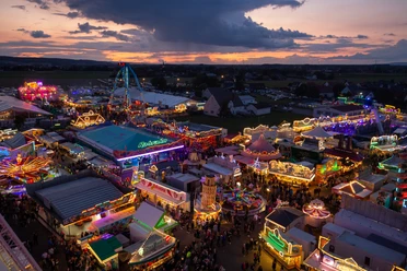 Lübbecke Blasheimer Markt Oliver Krato Abendlicher Jahrmarkt mit bunten Lichtern und Fahrgeschäften, umgeben von einer malerischen Landschaft.