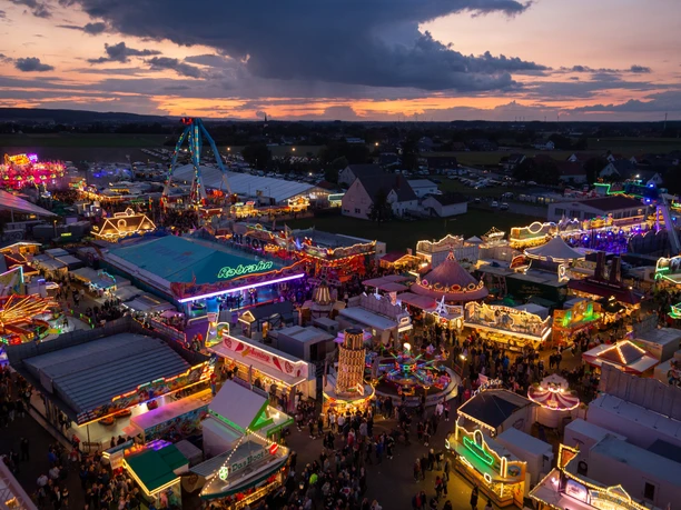 Lübbecke Blasheimer Markt Oliver Krato Abendlicher Jahrmarkt mit bunten Lichtern und Fahrgeschäften, umgeben von einer malerischen Landschaft.