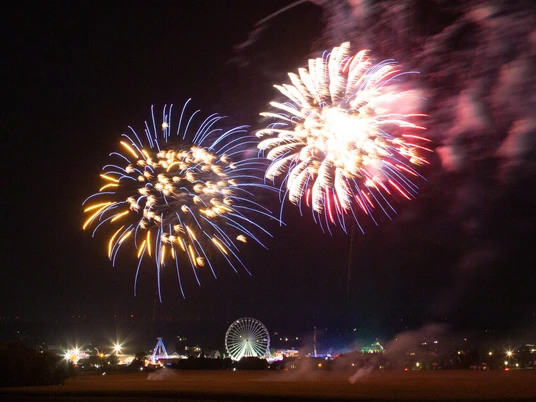 Leuchtendes Feuerwerk über einem Volksfest bei Nacht mit Riesenrad und bunten Lichtern im Hintergrund.