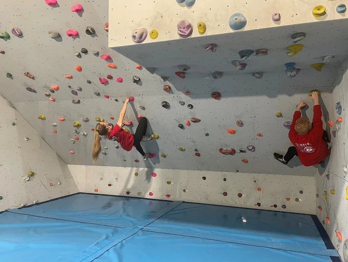 Zwei Personen klettern an farbigen Griffen einer Boulderwand in einer Indoor-Kletterhalle.