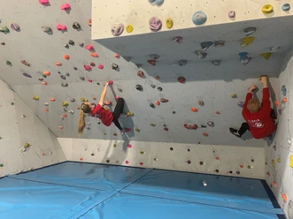 Kletterhalle Werlte.jpg Zwei Personen klettern an farbigen Griffen einer Boulderwand in einer Indoor-Kletterhalle.