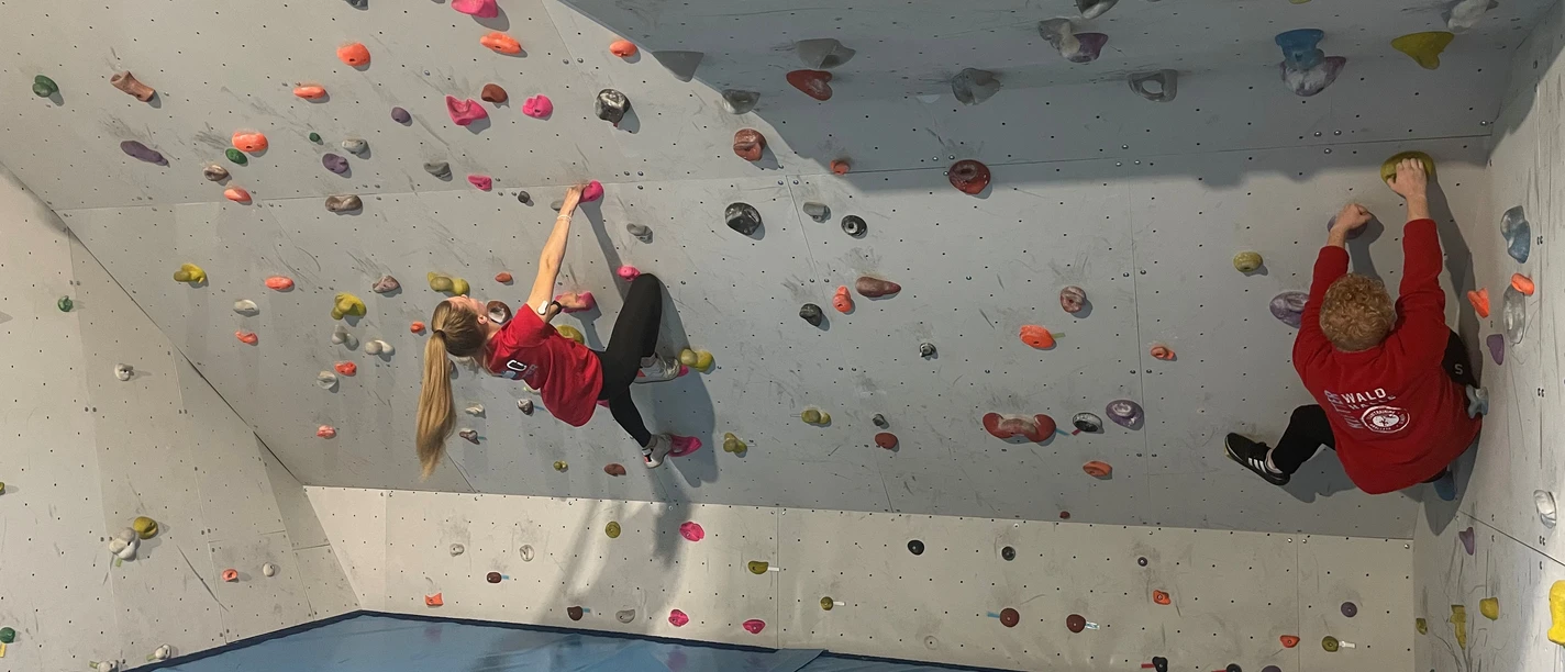 Kletterhalle Werlte.jpg Zwei Personen klettern an farbigen Griffen einer Boulderwand in einer Indoor-Kletterhalle.