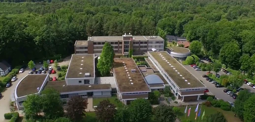 Titelbild Luftaufnahme eines großen, umgeben von Wald liegenden Gebäudekomplexes mit begrünten Flachdächern.