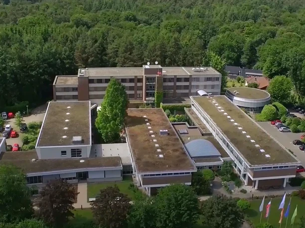 Titelbild Luftaufnahme eines großen, umgeben von Wald liegenden Gebäudekomplexes mit begrünten Flachdächern.