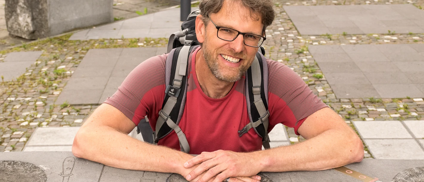 Stefan Hoene Ein Mann mit Brille und Rucksack lehnt lächelnd an einer Steinmauer mit eingravierten Symbolen.