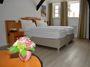 Doppelzimmer Superior 1.jpg Doppelzimmer mit modernen Möbeln, zwei Betten, großen Fenstern, Holzbalken an der Decke und Blumen.