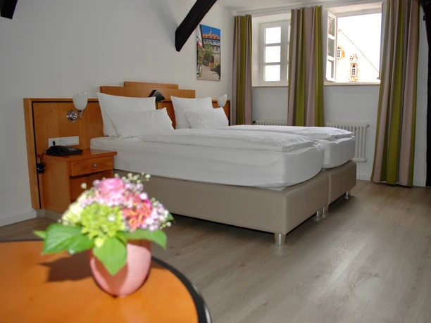 Doppelzimmer Superior 1.jpg Doppelzimmer mit modernen Möbeln, zwei Betten, großen Fenstern, Holzbalken an der Decke und Blumen.