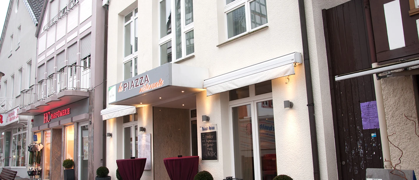 Ein italienisches Restaurant mit stilvoller Fassade, Markise und Stehtischen in einer Fußgängerzone.