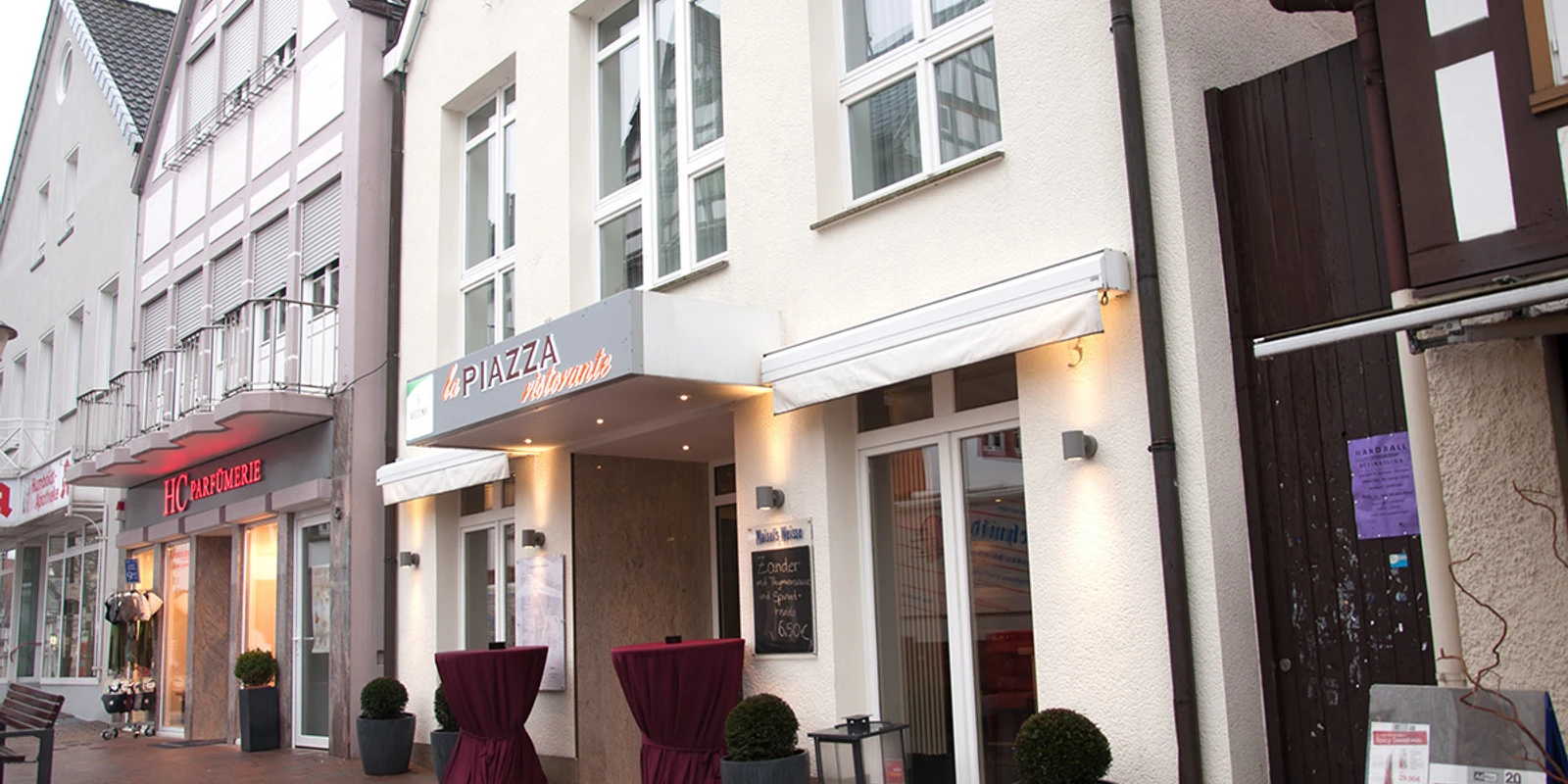 Blick von außen.jpg Ein italienisches Restaurant mit stilvoller Fassade, Markise und Stehtischen in einer Fußgängerzone.