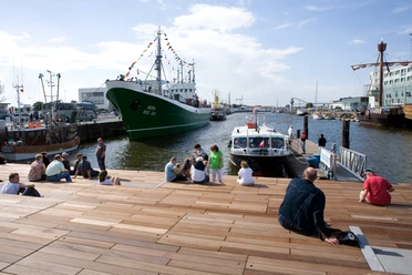 Menschen sitzen auf Holzstufen am Hafen, vor ihnen liegen Schiffe, darunter der Kutter GERA.