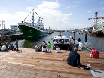 Schaufenster Fischereihafen Menschen sitzen auf Holzstufen am Hafen, vor ihnen liegen Schiffe, darunter der Kutter GERA.
