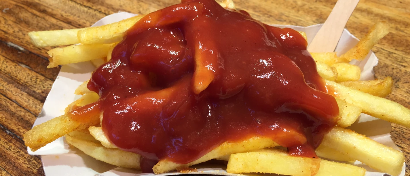 Pommes Pommes mit einer großzügigen Portion Ketchup auf einem weißen Pappteller, fotografiert auf einer Holzoberfläche.