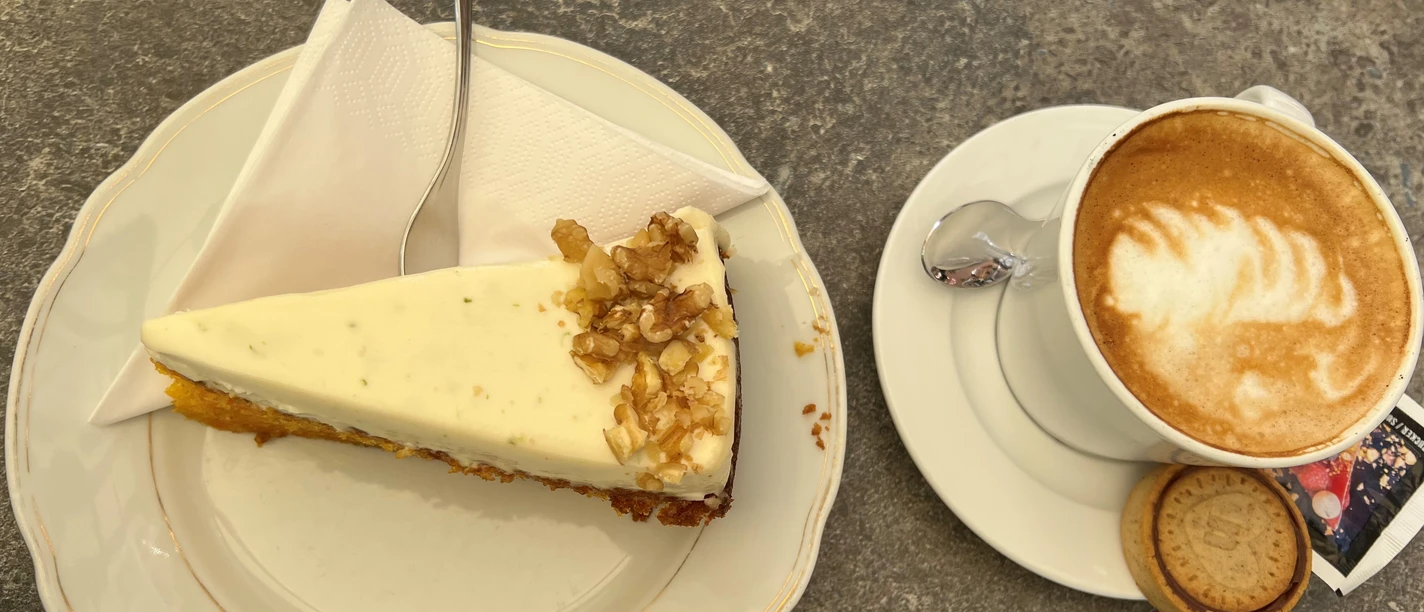 Kaffee_Kuchen Ein Stück Walnusskuchen mit Gabel auf Teller neben einer Tasse Cappuccino auf grauem Steintisch.