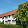 Fassade eines traditionellen Ferienhauses mit rotem Ziegeldach, umgeben von Grünen Büschen und Bäumen.