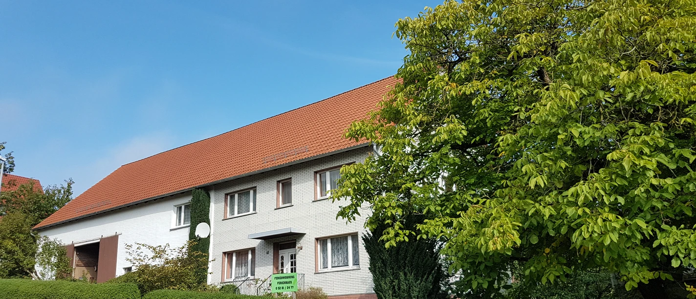 Fassade eines traditionellen Ferienhauses mit rotem Ziegeldach, umgeben von Grünen Büschen und Bäumen.