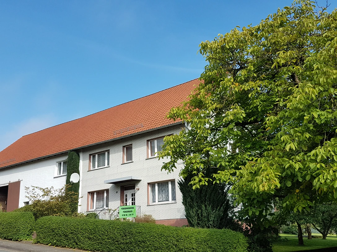 Ferienhaus Eschenbruch Fassade.jpg Fassade eines traditionellen Ferienhauses mit rotem Ziegeldach, umgeben von Grünen Büschen und Bäumen.