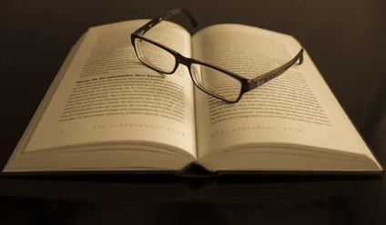 Ein offenes Buch mit Lesebrille aufgeschlagen auf einem Tisch in gedämpfter Beleuchtung.