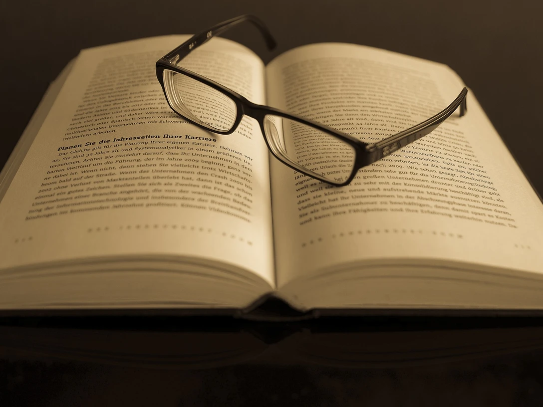 Ein offenes Buch mit Lesebrille aufgeschlagen auf einem Tisch in gedämpfter Beleuchtung.