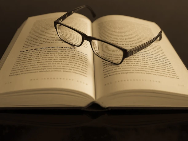 Ein offenes Buch mit Lesebrille aufgeschlagen auf einem Tisch in gedämpfter Beleuchtung.