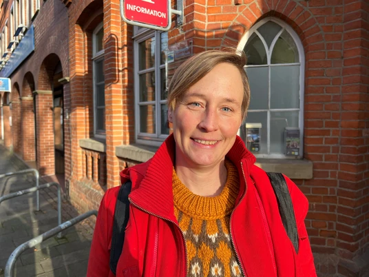Frau Matissek Porträt einer lächelnden Frau vor einem Backsteingebäude mit gotischen Fenstern und sonnigem Wetter.
